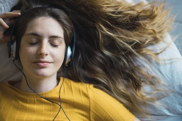an overhead portrait of a young adult lying on bed listening to music with headphones