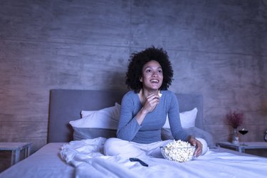 Woman smiling watching TV in bed with bowl of popcorn