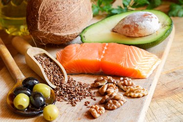 Foods high in healthy fats, including avocado, salmon, olives, nuts and seeds, on wooden board