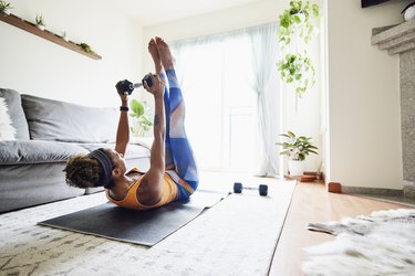 Person in orange tank top and blue leggings holding dumbbell and doing a 10-minute chest and arms workout lying down