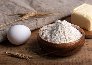 Flour in a wooden bowl, butter and an egg on a wooden table.