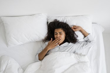 an overhead photo of a person yawning while lying in bed with white sheets and pillowcases