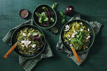 top view of two bowls of green vegetable pasta and a smaller bowl of green salad on a green tablecloth