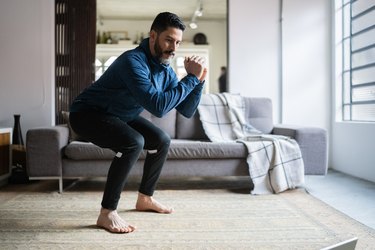 Mature man with dark hair and beard doing a squat exercise in his living room
