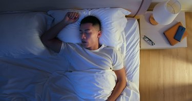 overhead view of a person with short brown hair and a white t-shirt sleeping in bed with arm above their head