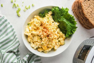 Instant Pot Egg Salad in a white bowl with lettuce leave next to slices of whole grain bread.
